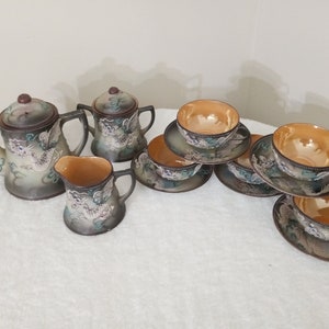 May include: A vintage Japanese tea set with a dragon design. The set includes a teapot, creamer, and four teacups and saucers. The teacups and saucers are decorated with a floral pattern.