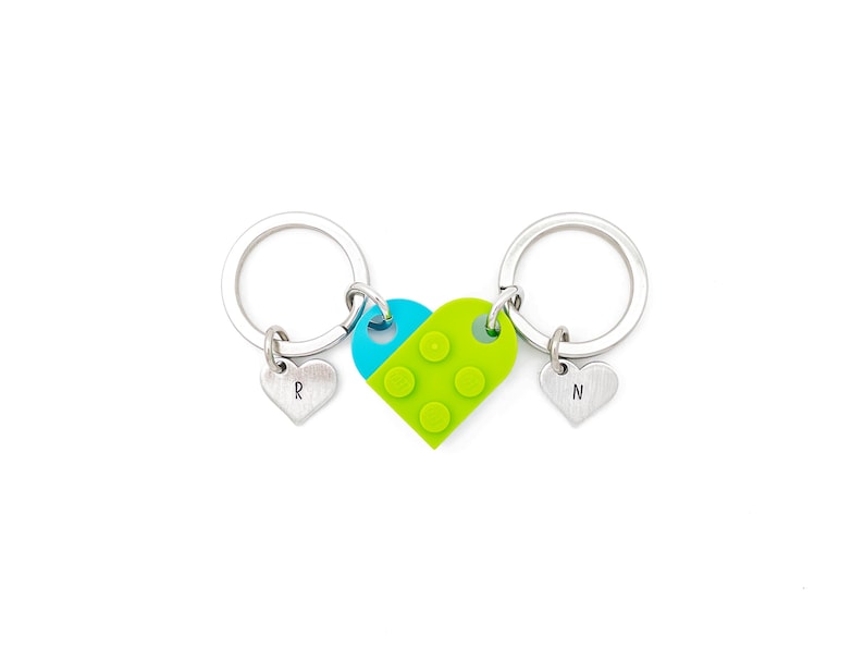 Puede incluir: Dos llaveros plateados con dijes en forma de coraz&oacute;n. Los dijes est&aacute;n conectados por un dije en forma de coraz&oacute;n verde y azul con un dise&ntilde;o tipo Lego. Los dijes est&aacute;n grabados con las letras "R" y "N".