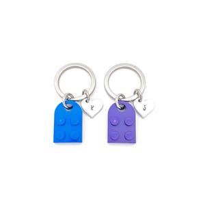 Puede incluir: Dos llaveros con anillos plateados, dijes de bloques de Lego azul y morado, y dijes de coraz&oacute;n plateados con las letras "K" y "S" grabadas.