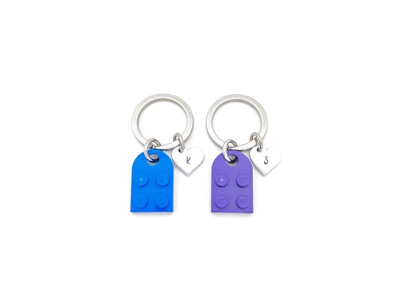 Puede incluir: Dos llaveros con anillos plateados, dijes de bloques de Lego azul y morado, y dijes de coraz&oacute;n plateados con las letras "K" y "S" grabadas.
