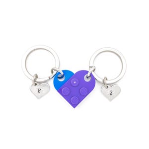 Puede incluir: Dos llaveros plateados con dijes en forma de coraz&oacute;n, cada uno con una letra grabada. Los llaveros est&aacute;n conectados por un dije en forma de coraz&oacute;n morado y azul que se asemeja a un ladrillo Lego.