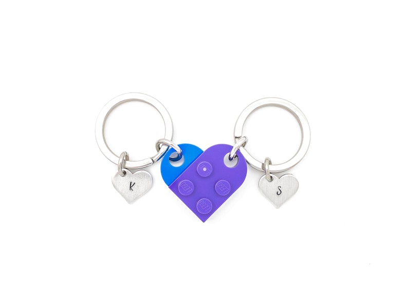 Puede incluir: Dos llaveros plateados con dijes en forma de coraz&oacute;n, cada uno con una letra grabada. Los llaveros est&aacute;n conectados por un dije en forma de coraz&oacute;n morado y azul que se asemeja a un ladrillo Lego.