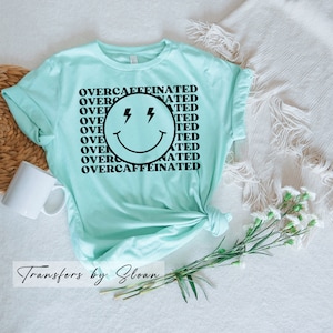 May include: A light teal t-shirt with the words "OVERCAFFEINATED" in a repeating pattern around a smiley face with lightning bolt eyes. The shirt is knotted at the bottom and has rolled-up sleeves. A white mug and flowers are in the background.