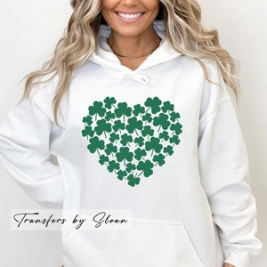 May include: White hoodie featuring a heart design made of green shamrocks. The hoodie has a drawstring hood and a front pocket. The text "Transfers by Sloan" is printed at the bottom left of the image.