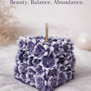 May include: A square candle with a floral design in shades of purple and white. The candle has a wick and the text "Beauty. Balance. Abundance.". This decorative candle is suitable for home decor.