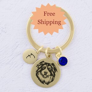 May include: Gold keychain with a round charm featuring a black and white silhouette of an Australian Shepherd dog. The charm also has a small gold charm with the letter "J" and a blue crystal charm.