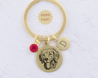 May include: Gold keychain with a round charm featuring a black and white illustration of a dog's head. The charm is attached to a gold split ring with a small red crystal charm and a round charm with the letter "D" engraved on it.