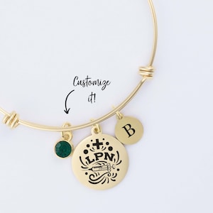 May include: Gold bangle bracelet with a charm that says "LPN" with a cross and a syringe. The bracelet also has a green gemstone charm and a charm with the letter "B".
