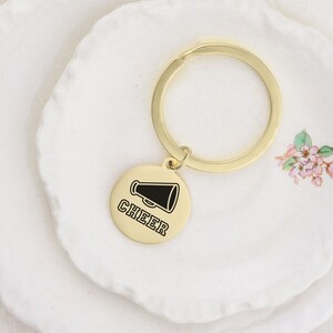 Puede incluir: Llavero dorado con un dije redondo que presenta un meg&aacute;fono negro y la palabra "CHEER" en texto negro.