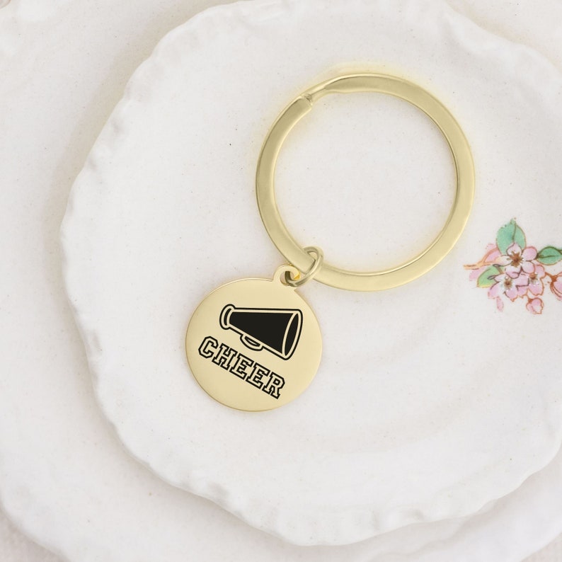 Puede incluir: Llavero dorado con un dije redondo que presenta un meg&aacute;fono negro y la palabra "CHEER" en texto negro.