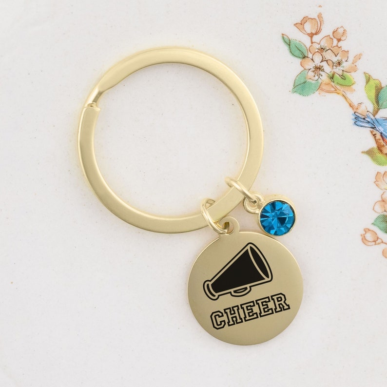 Puede incluir: Llavero dorado con un dije redondo que presenta un meg&aacute;fono negro y la palabra "CHEER". Un peque&ntilde;o dije de cristal azul cuelga del anillo del llavero.