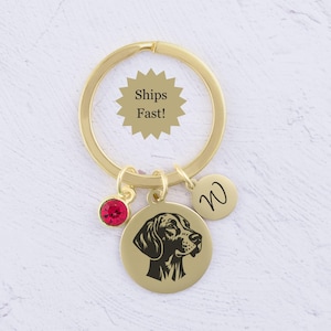 May include: A gold-toned metal keychain featuring a circular charm engraved with a dog profile, a smaller circular charm engraved with the letter W, and a small round red crystal charm attached to a gold split ring.