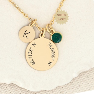 May include: Gold necklace with a round charm engraved with the coordinates 40.71128° N 74.0060° W, a small round charm with the letter K, and a green crystal charm.