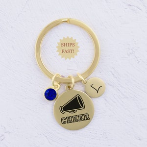 Puede incluir: Llavero dorado con un dije de cristal azul, un peque&ntilde;o dije circular con la letra V y un dije circular m&aacute;s grande con un meg&aacute;fono negro y la palabra "CHEER" debajo.