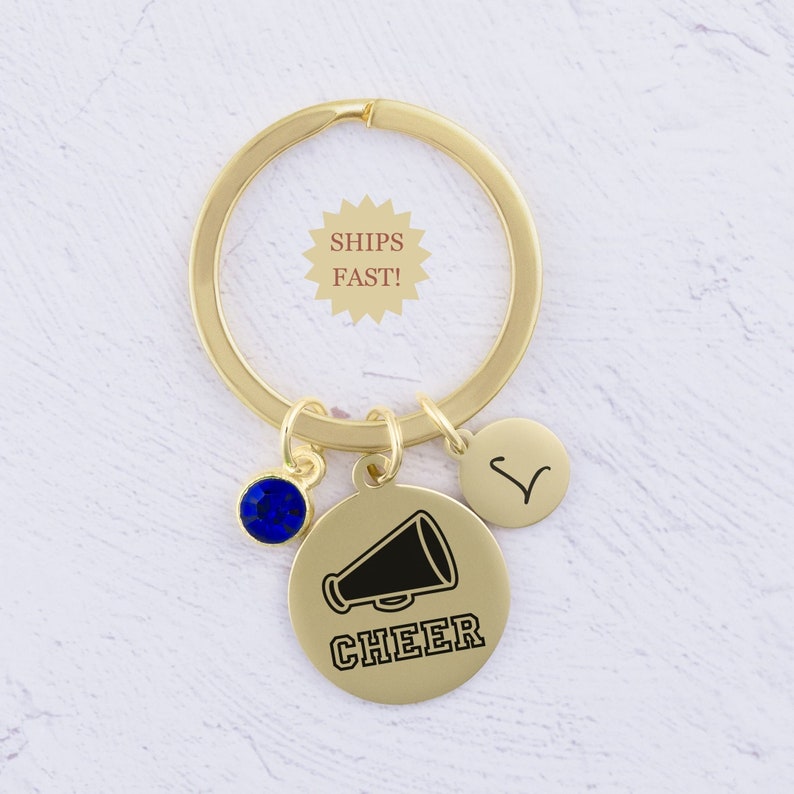 Puede incluir: Llavero dorado con un dije de cristal azul, un peque&ntilde;o dije circular con la letra V y un dije circular m&aacute;s grande con un meg&aacute;fono negro y la palabra "CHEER" debajo.