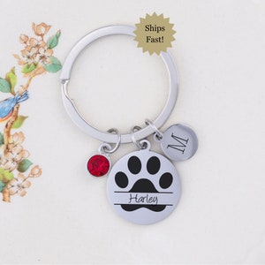 May include: Silver keychain with a black paw print charm, a red crystal charm, and a silver charm with the letter M. The paw print charm has the name Harley engraved on it.