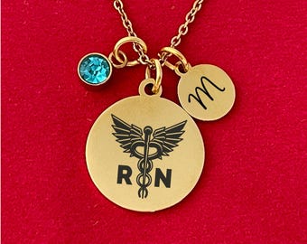 May include: Gold necklace with a blue crystal charm, a gold charm with a medical caduceus symbol and the letters "RN", and a gold charm with the letter "M".