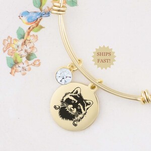 May include: Gold bangle bracelet with a charm featuring a black and white raccoon illustration. The bracelet also has a clear crystal charm.