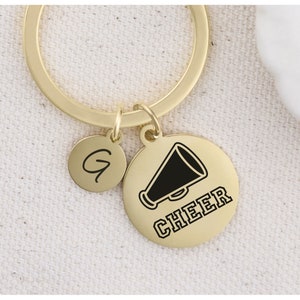 Puede incluir: Llavero dorado con un dije redondo que presenta un meg&aacute;fono negro y la palabra "CHEER". Un dije redondo m&aacute;s peque&ntilde;o con la letra "G" est&aacute; unido al llavero.