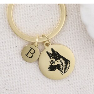 May include: Gold-toned keychain with a round charm featuring a black and white silhouette of a German Shepherd's head. A smaller round charm with the letter "B" is attached to the keychain.