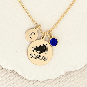 May include: Gold necklace with a charm that says "CHEER" and a megaphone graphic. There is also a small blue crystal charm and a charm with the letter "L" on it.