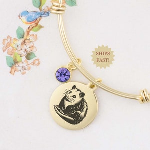 May include: Gold bangle bracelet with a charm featuring a black and white opossum illustration. The bracelet also has a purple crystal charm.