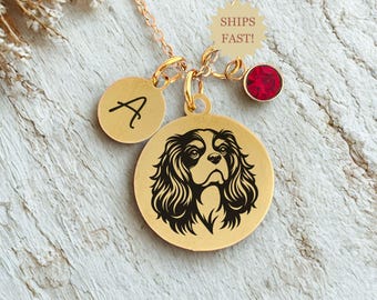 May include: Gold-colored pendant necklace featuring a Cavalier King Charles Spaniel design. The necklace includes a round charm with the letter "A", a red gemstone, and a charm that says "SHIPS FAST!"