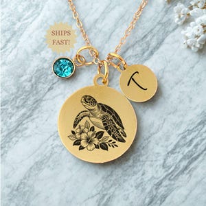 May include: Gold-colored necklace featuring a pendant with a sea turtle and floral design. The necklace includes a teal-colored gemstone and a round charm with a letter. The text "SHIPS FAST!" is also visible.