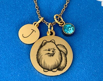May include: Gold necklace with three charms: a round charm with the letter J, a round charm with a black and white silhouette of a Pomeranian dog, and a small round charm with a light blue crystal.