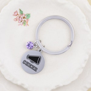 Puede incluir: Un llavero plateado con un dije de cristal morado y un dije redondo plateado con la palabra "CHEER" y un gr&aacute;fico de meg&aacute;fono negro.