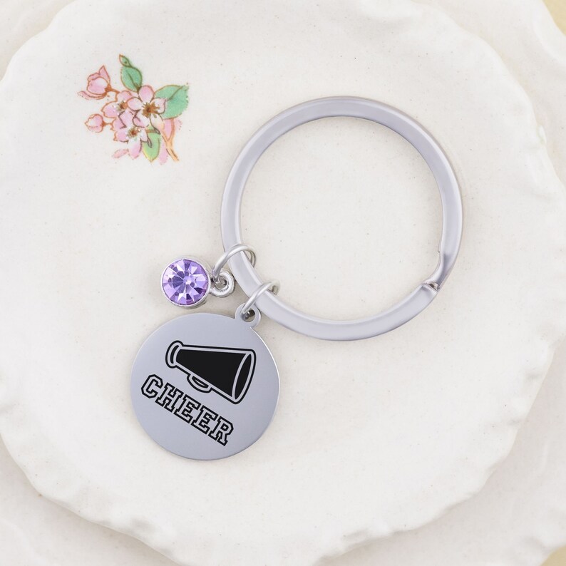 Puede incluir: Un llavero plateado con un dije de cristal morado y un dije redondo plateado con la palabra "CHEER" y un gr&aacute;fico de meg&aacute;fono negro.