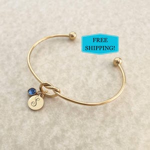 May include: Gold knot bangle bracelet with a small charm featuring a blue crystal and a gold disc with the letter 'S' engraved on it.