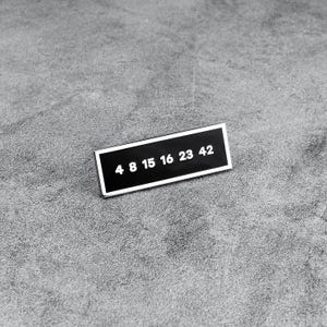 Puede incluir: Pin de esmalte rectangular en blanco y negro con los números "4 8 15 16 23 42" en blanco. El pin está sobre un fondo gris texturizado.