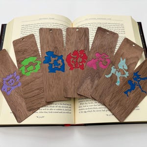 May include: Six wooden bookmarks with colorful designs rest on an open book. Each bookmark features a different design in purple, green, blue, pink, and teal. The bookmarks are made of dark wood and have a hole at the top.