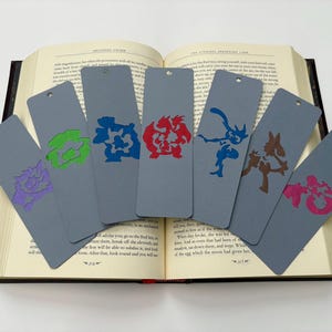 May include: Six gray bookmarks with colorful character silhouettes. Each bookmark has a different character in purple, green, blue, red, blue, brown, and pink. The bookmarks are resting on an open book.