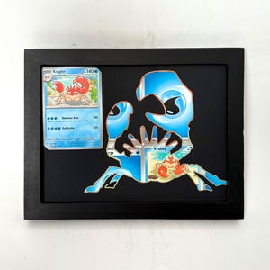 May include: Framed artwork featuring a blue and white Krabby design with a matching Kingler card. The artwork is set against a black background within a black frame, creating a striking contrast.