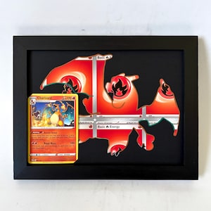 May include: A black rectangular shadow box frame containing a Charizard trading card on the left and a large, stylized silhouette of the same creature crafted from red and white fire energy cards on a black background.
