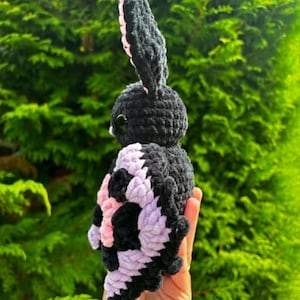 May include: A handmade crochet plush toy shaped like a moth with a black body and large, textured wings featuring a pattern of black and light purple yarn. The creature has prominent, upright black antennae and small, dark, glossy eyes on its head.