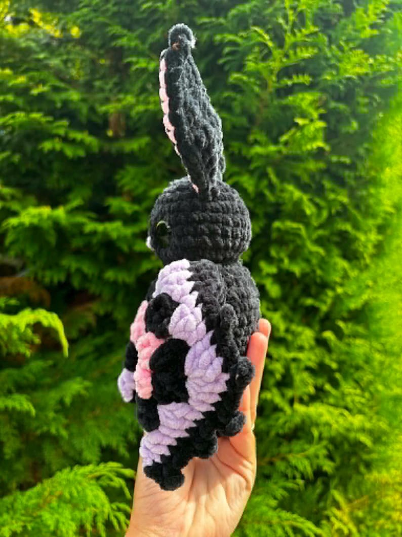 May include: A handmade crochet plush toy shaped like a moth with a black body and large, textured wings featuring a pattern of black and light purple yarn. The creature has prominent, upright black antennae and small, dark, glossy eyes on its head.