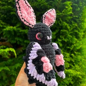 May include: A crocheted bat plush toy featuring a black body, large pointed ears with pink inner detailing, and wings accented with light purple and pink yarn. The creature has round, glossy black eyes with a subtle red reflection.