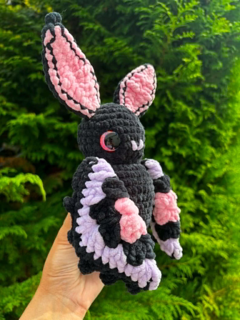 May include: A crocheted bat plush toy featuring a black body, large pointed ears with pink inner detailing, and wings accented with light purple and pink yarn. The creature has round, glossy black eyes with a subtle red reflection.