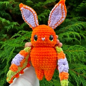May include: A plush crochet creature featuring a bright orange body, large pointed ears with light purple centres, and wide black eyes. The creature has outstretched wings patterned with orange, light purple, and light green yarn segments.
