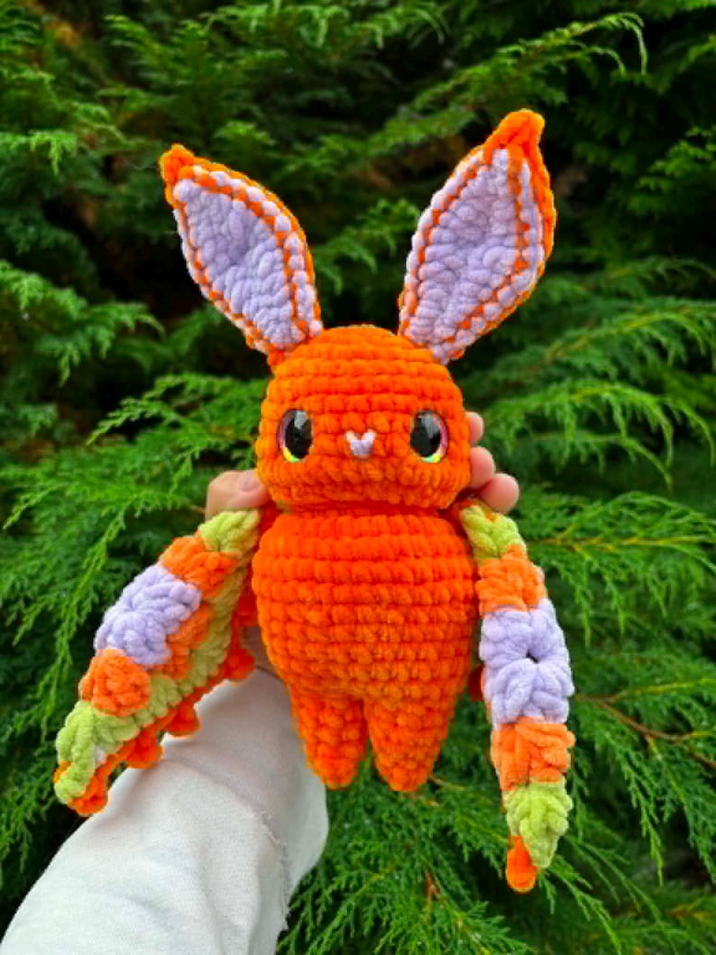 May include: A plush crochet creature featuring a bright orange body, large pointed ears with light purple centres, and wide black eyes. The creature has outstretched wings patterned with orange, light purple, and light green yarn segments.