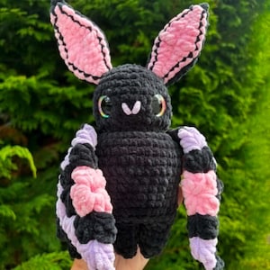 May include: A handmade crochet plush toy shaped like a bat with a black body, large pointed ears with pink inner sections, and wide wings featuring alternating stripes of black, pink, and light purple yarn. The bat has large, glossy, dark eyes.