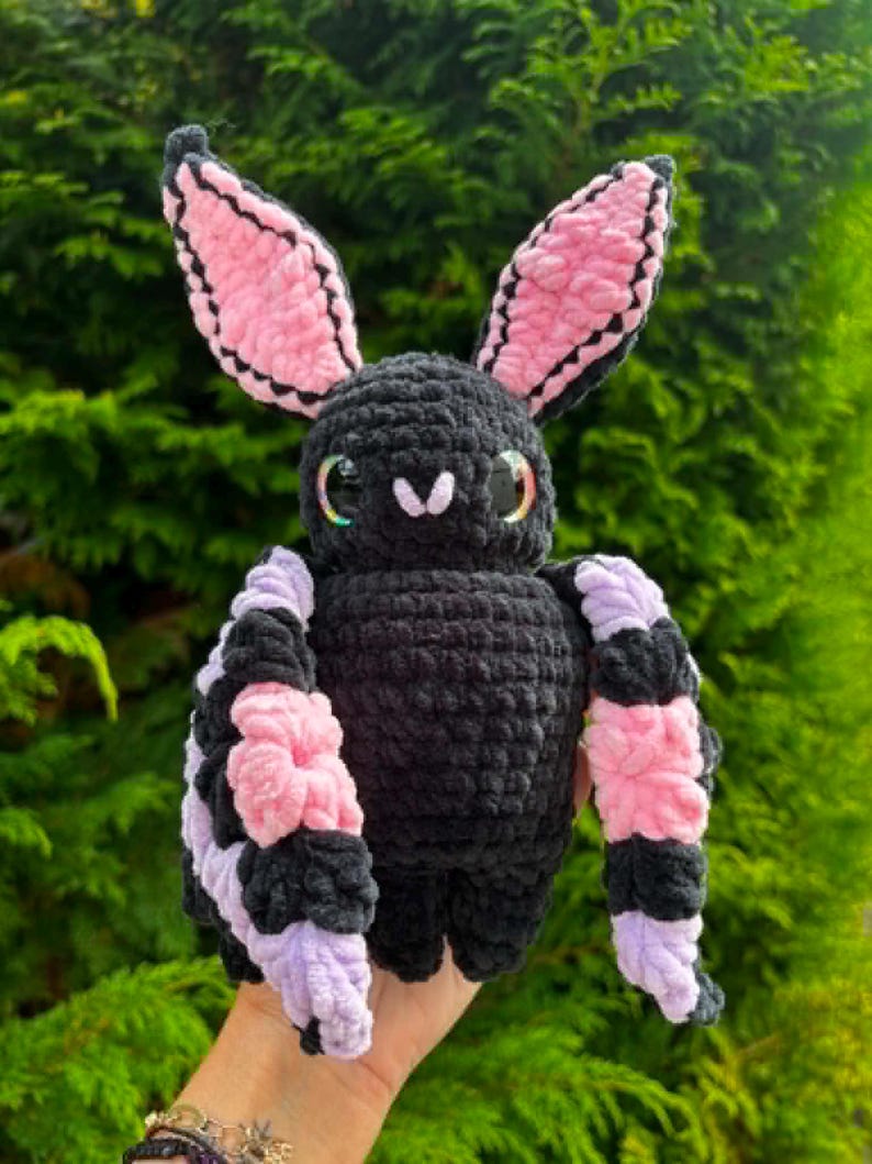 May include: A handmade crochet plush toy shaped like a bat with a black body, large pointed ears with pink inner sections, and wide wings featuring alternating stripes of black, pink, and light purple yarn. The bat has large, glossy, dark eyes.