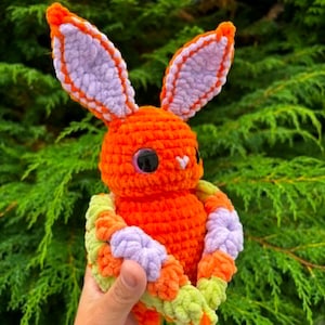 May include: A handmade crochet plush toy shaped like a rabbit with bright orange yarn, featuring large lavender inner ears, dark glossy eyes, and a small white nose. The rabbit is wrapped in a textured blanket made of orange, lavender, and light green yarn.