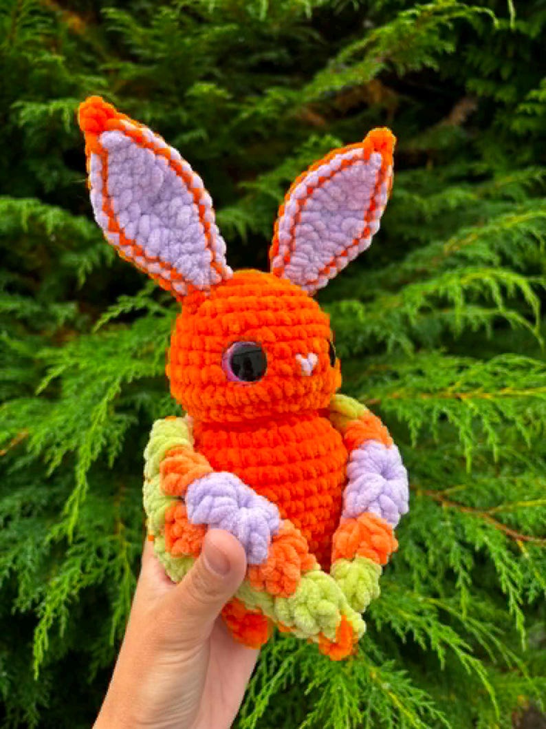 May include: A handmade crochet plush toy shaped like a rabbit with bright orange yarn, featuring large lavender inner ears, dark glossy eyes, and a small white nose. The rabbit is wrapped in a textured blanket made of orange, lavender, and light green yarn.