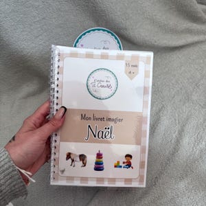 Peut inclure: Un livre à spirales avec un motif à carreaux beiges et blancs. La couverture présente le texte "Mon livret imagier Noël" et des illustrations d'un poney, d'une pile d'anneaux et d'un enfant jouant avec des blocs. Le livre est étiqueté "15 mois et +."