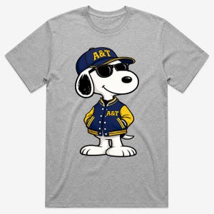 Op de afbeelding: Een grijs T-shirt met een cartoonhond die een marineblauwe baseballcap en jasje met gele accenten draagt. Op de pet en jas staan de letters "A&T" in goud. De hond draagt ook een zwarte zonnebril.