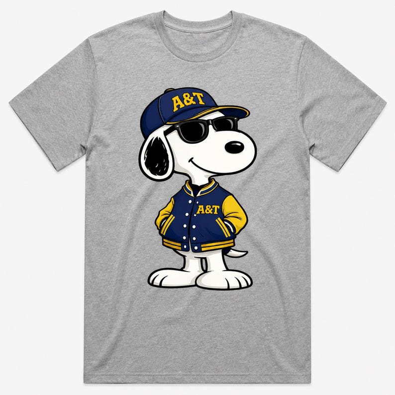 Op de afbeelding: Een grijs T-shirt met een cartoonhond die een marineblauwe baseballcap en jasje met gele accenten draagt. Op de pet en jas staan de letters "A&T" in goud. De hond draagt ook een zwarte zonnebril.